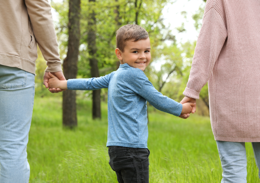 Young boy holding hands with parents in a lush green park.