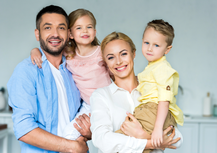 A happy family of four with two young children smiling at home.