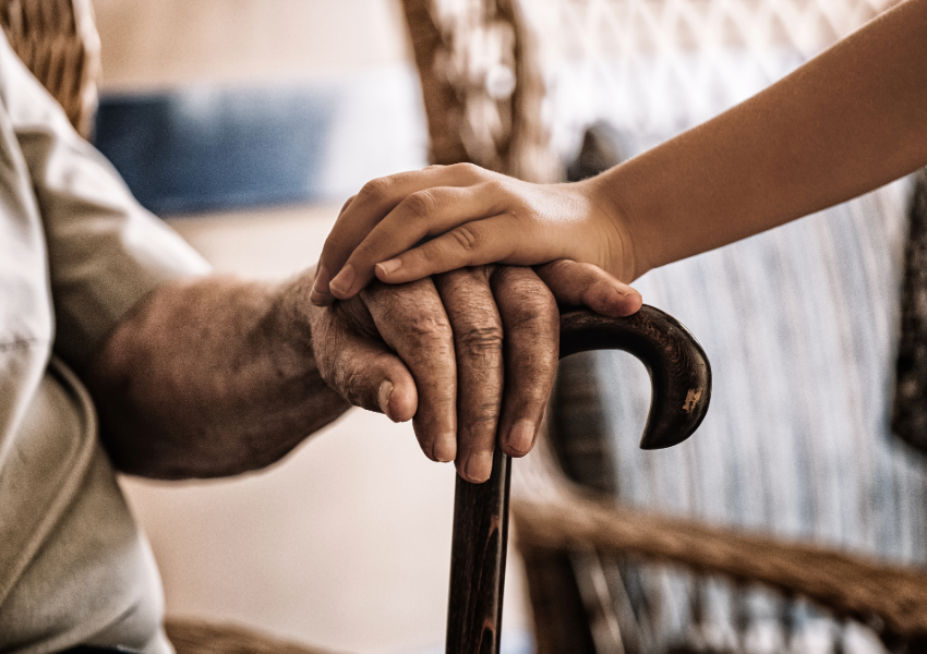 A young hand holding an elderly hand resting on a cane.