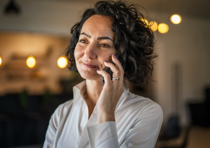 A thoughtful woman smiling while talking on the phone in a well-lit room.