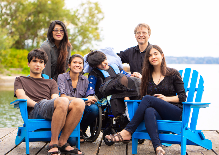 Diverse family smiling by the lake, one in a wheelchair.